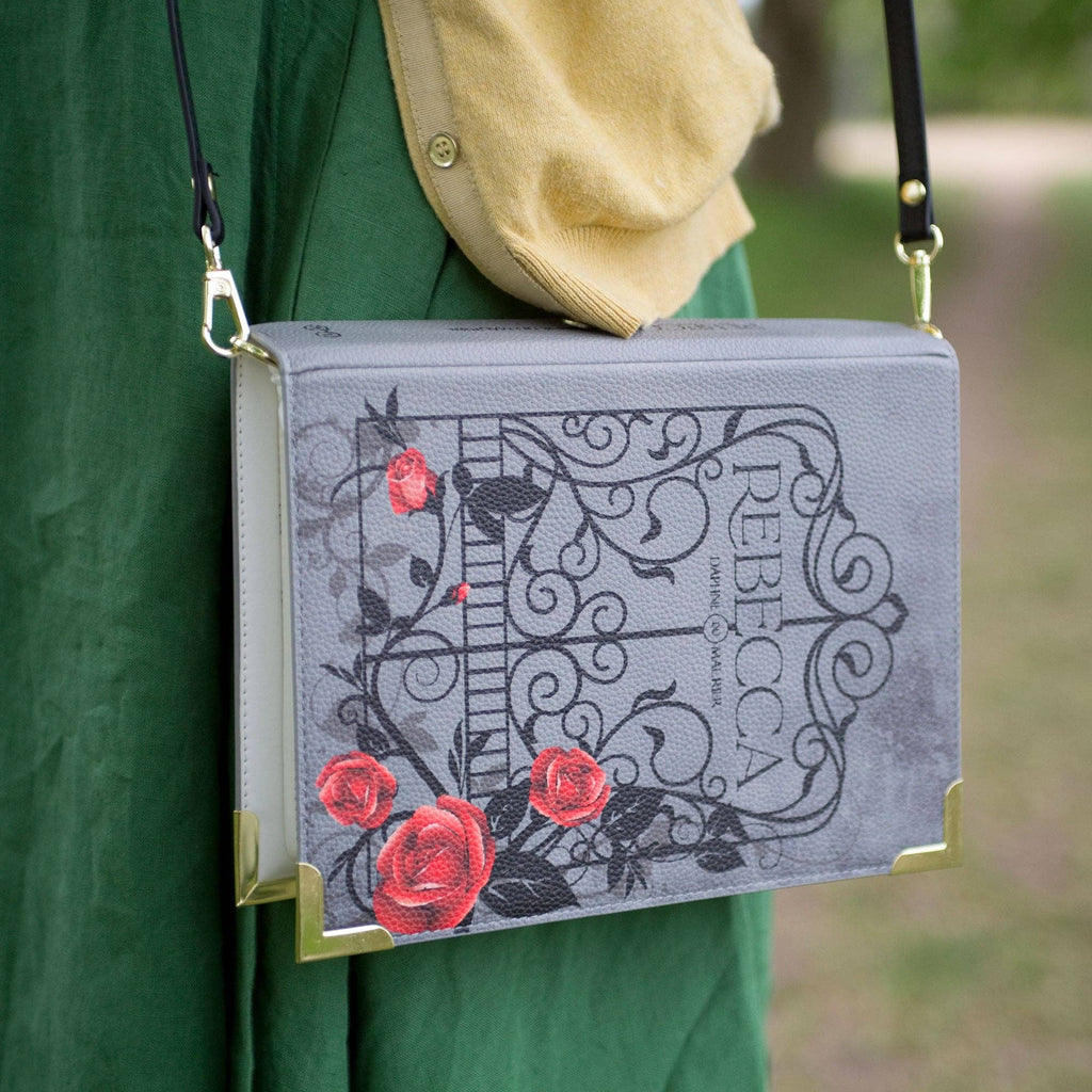 Book-shaped handbag with decorative design and 'Rebecca' text, held by a person outdoors.