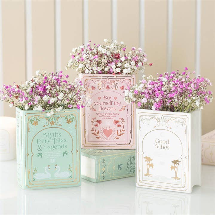 Three decorative planters with flowers inside, each on a book titled 'Myths, Fairy Tales & Legends', 'Buy Yourself the Flowers', and 'Good Vibes'.