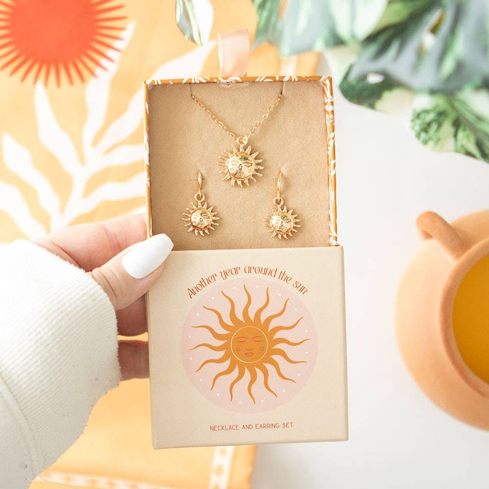 Gold necklace and earrings set in a decorative box with sun design, held by a hand.