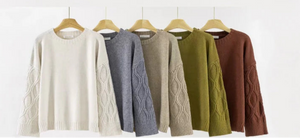 Row of five cable knit sweaters in different colors on hangers against a white background