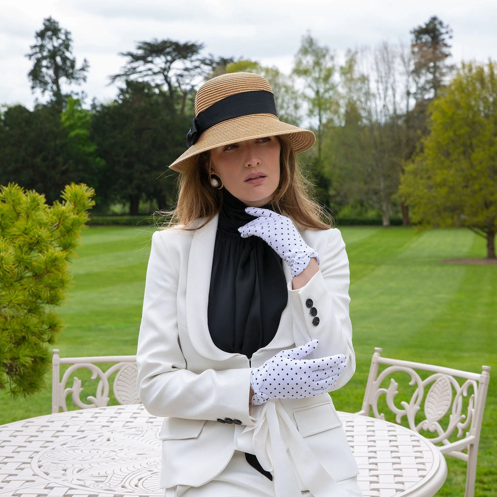 Woman in formal attire with a hat and gloves sitting outdoors on a lawn.